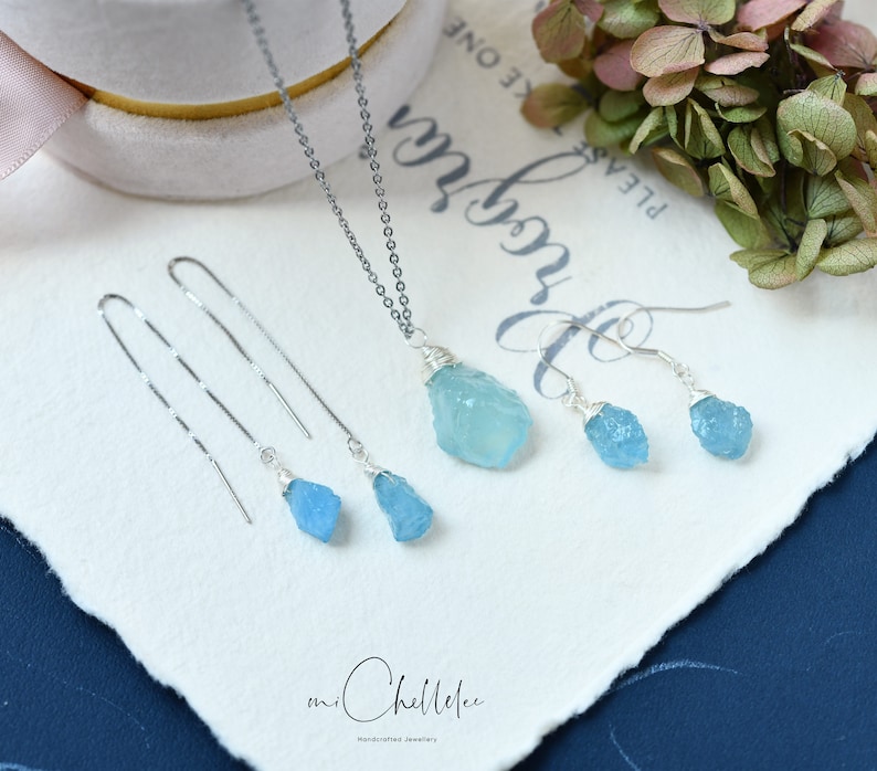 May include: A set of aquamarine gemstone jewelry featuring a necklace and earrings. The necklace has a silver chain and a pendant with a raw aquamarine stone. The earrings are silver with raw aquamarine stones.