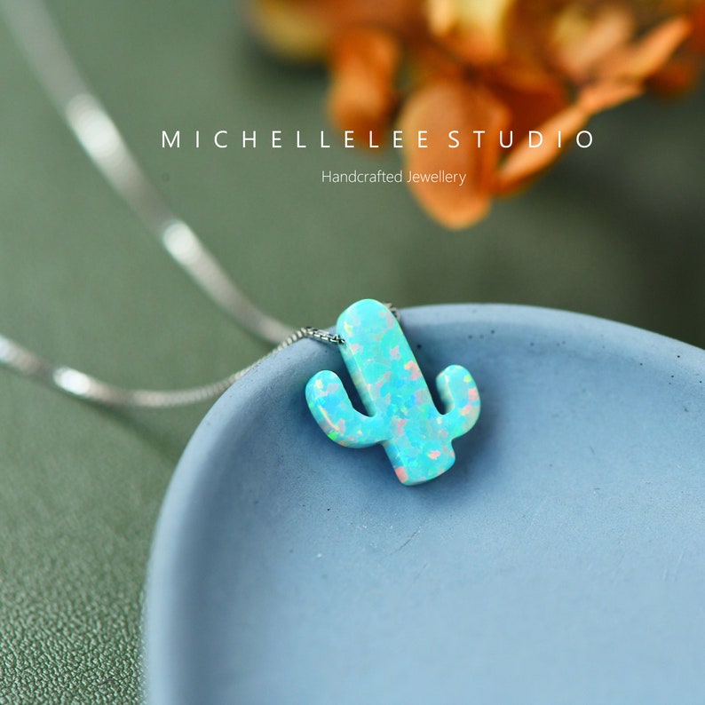 Op de afbeelding: Een zilveren ketting met een blauwe opaal cactus hanger. De hanger is lichtblauw met witte en roze accenten. De ketting ligt op een blauw oppervlak. Handgemaakt sieraden.