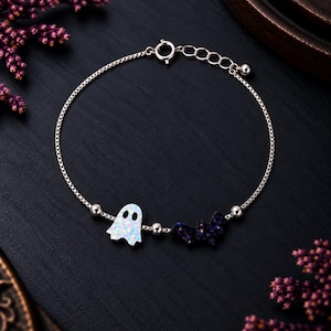 Op de afbeelding: Een zilveren kettingarmband met twee bedels: een witte iriserende geest en een donkerpaarse glitterende vleermuis. Het armbandje heeft een fijne boxketting, kleine zilveren kraaltjes tussen de bedels en een ronde veerringsluiting.