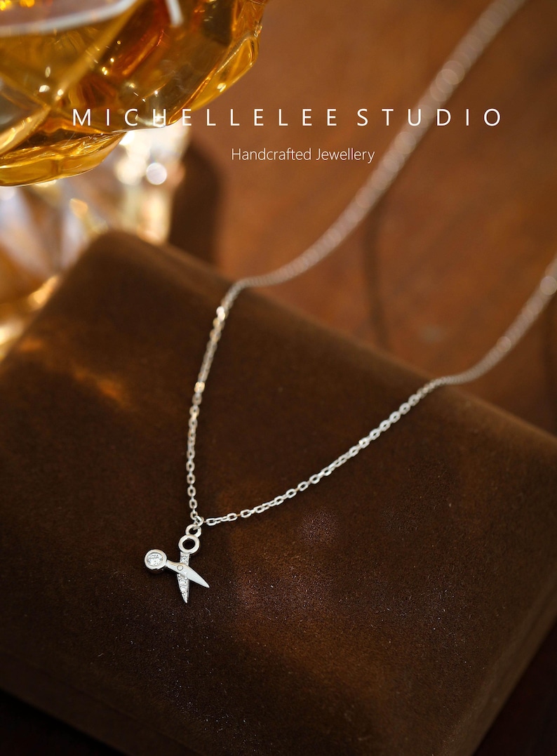 May include: A silver necklace with a small, delicate pendant shaped like a pair of scissors with a single rhinestone on the ring. The necklace is on a brown velvet surface.  Michelle Lee Studio. Handcrafted Jewellery.