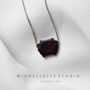 May include: A silver chain necklace with a red and black pendant. The pendant is a small, rectangular shape with a glittery finish. The necklace is on a white background.
