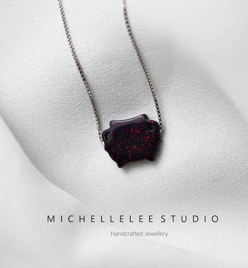 May include: A silver chain necklace with a red and black pendant. The pendant is a small, rectangular shape with a glittery finish. The necklace is on a white background.