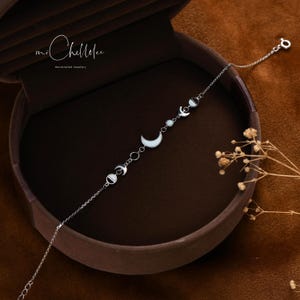 Peut inclure: Un bracelet en argent avec une cha&icirc;ne d&eacute;licate et des breloques en forme de lune et de croissant de lune. Le bracelet est pr&eacute;sent&eacute; dans une bo&icirc;te brune avec le texte "mi Chillee Handmade Jewelry".