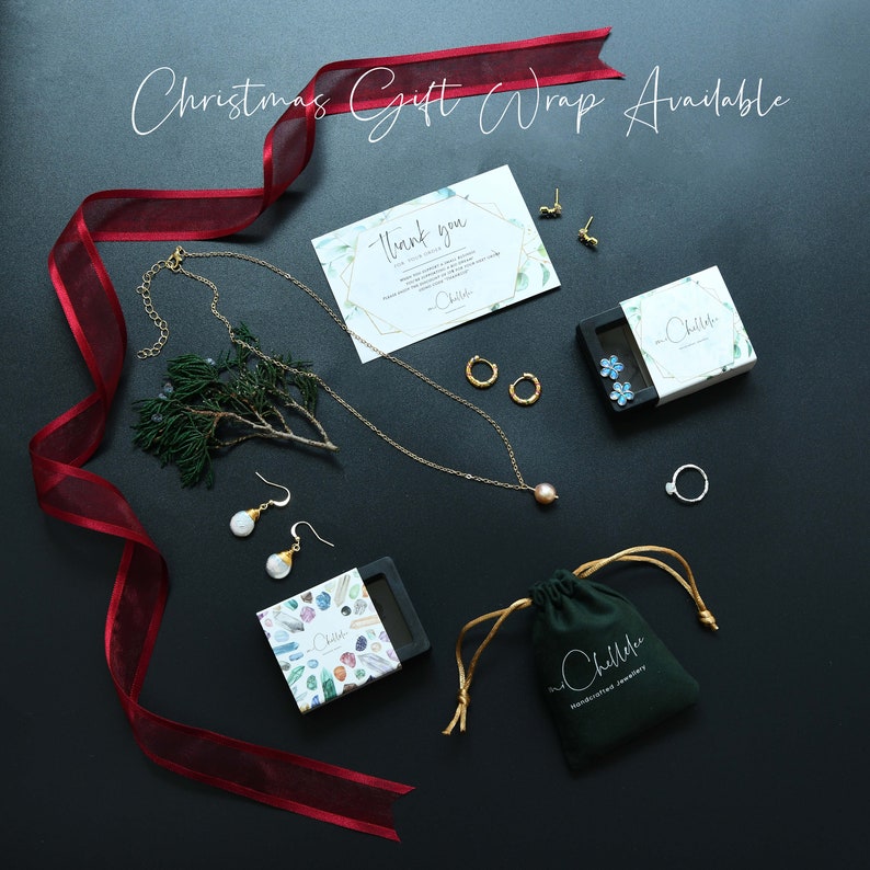 May include: A flat lay image of jewelry and gift wrapping supplies. A gold necklace with a pearl pendant, a pair of pearl earrings, a pair of gold hoop earrings, a ring, and a small box with a blue flower design are arranged on a black surface. A red ribbon and a small green plant are also in the image. The text "Christmas Gift Wrap Available" is written in white at the top of the image.