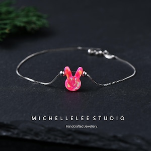 May include: A delicate silver bracelet featuring a pink opal bunny charm. The bunny charm has a textured surface and is accented with small silver beads. The bracelet is displayed on a dark surface. The text "MICHELLELEE STUDIO Handcrafted Jewellery" is at the bottom.