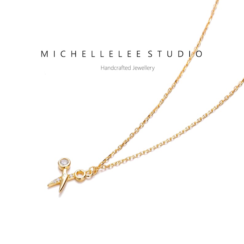 May include: A gold chain necklace with a small gold pendant shaped like a pair of scissors with a single clear gemstone in the center.
