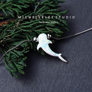 Può includere: Una collana d'argento con un ciondolo a forma di delfino. Il ciondolo è bianco con accenti iridescenti blu e rosa. La collana è esposta su una superficie scura con fogliame verde sullo sfondo. Il testo "MICHELLELEE STUDIO Handcrafted Jewellery" è visibile.