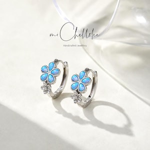 May include: A pair of silver hoop earrings featuring a five-petal flower design with iridescent blue opal-like inlays and a smaller plain silver flower accent. The earrings are marked with 925 and displayed below the text mi Chellelee Handcrafted Jewellery.