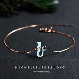Puede incluir: Una delicada pulsera de oro rosa con un dije de sirena con acabado opalino. El dije est&aacute; centrado en la cadena, flanqueado por dos peque&ntilde;as cuentas redondas. La pulsera se muestra sobre una superficie oscura. El texto "MICHELLELEE STUDIO Handcrafted Jewellery" es visible.