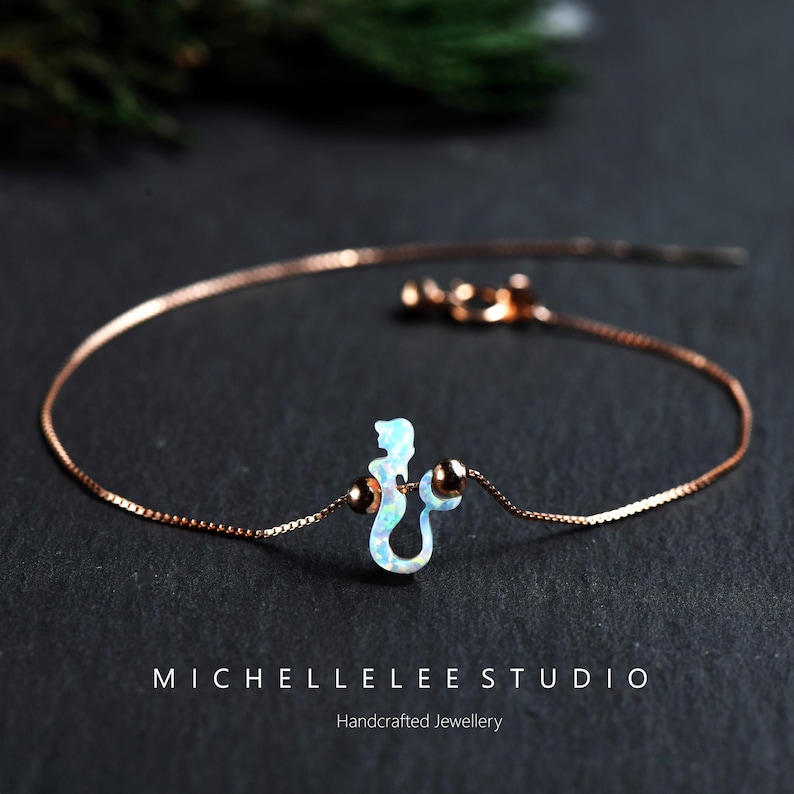 Puede incluir: Una delicada pulsera de oro rosa con un dije de sirena con acabado opalino. El dije est&aacute; centrado en la cadena, flanqueado por dos peque&ntilde;as cuentas redondas. La pulsera se muestra sobre una superficie oscura. El texto "MICHELLELEE STUDIO Handcrafted Jewellery" es visible.