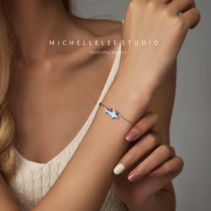 May include: A silver bracelet with a light blue shark charm. The bracelet is on a woman's wrist. The text "MICHELLELEE STUDIO" and "Handcrafted Jewellery" is visible in the image.