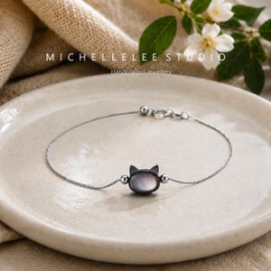 May include: A delicate silver-toned chain bracelet featuring a central cat-shaped charm made of iridescent dark shell material. The charm is flanked by two small silver-toned beads on a thin, flexible chain with a secure lobster claw clasp closure.