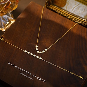 May include: A gold necklace and bracelet set with white pearl accents, displayed on a dark wooden surface. The jewelry is delicate and elegant, with a minimalist design. The brand name "MICHELLELEE STUDIO" is visible.