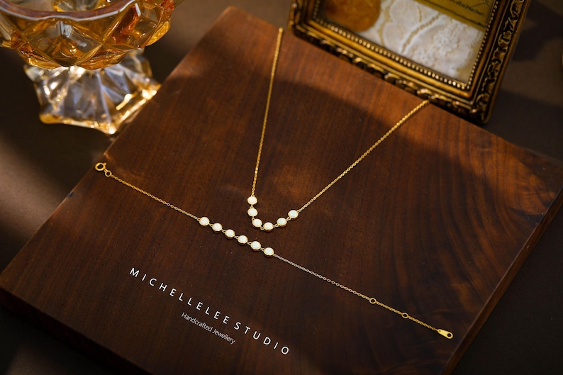 May include: A gold necklace and bracelet set with white pearl accents, displayed on a dark wooden surface. The jewelry is delicate and elegant, with a minimalist design. The brand name "MICHELLELEE STUDIO" is visible.