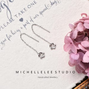 Könnte beinhalten: Silberne Kettenohrringe mit kleinen Blumenanhängern. Die Ohrringe werden auf einer weißen Oberfläche mit dem Text "PLEASE TAKE ONE" und "MICHELLELEE STUDIO Handcrafted Jewellery" präsentiert. Rosa Blüten sind im Hintergrund.