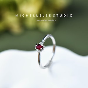 Peut inclure: Une bague en argent avec une pierre ovale rouge et deux petites pierres claires. La bague est sur une surface blanche. MICHELLELEESTUDIO Bijoux artisanaux