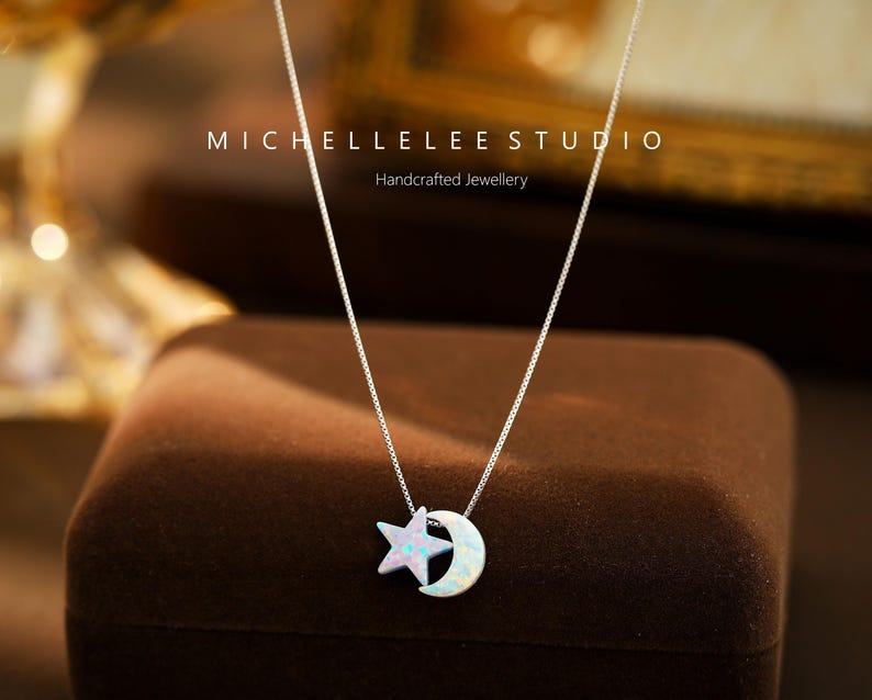 May include: A silver chain necklace with a star and crescent moon pendant made of white opal. The pendant is on a brown velvet surface. The text "MICHELLELEE STUDIO Handcrafted Jewellery" is visible above the pendant.