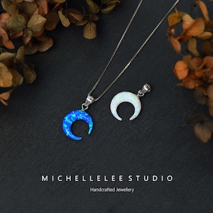 May include: Two silver necklaces with crescent moon pendants. One pendant is blue opal and the other is white opal. The necklaces are on a black background with brown leaves.