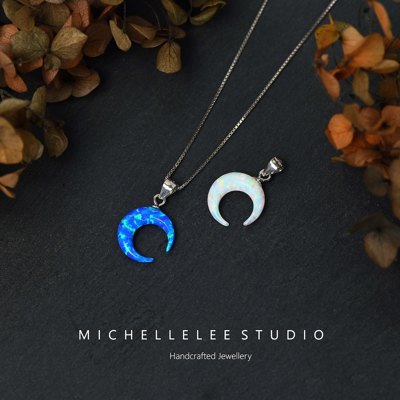 May include: Two silver necklaces with crescent moon pendants. One pendant is blue opal and the other is white opal. The necklaces are on a black background with brown leaves.