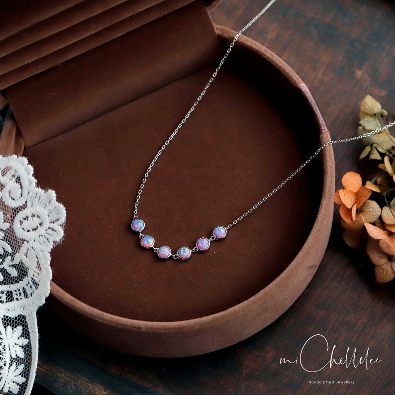 May include: A delicate silver necklace featuring seven round, iridescent opal stones. The opals display a range of pink and blue hues. The necklace is displayed in a brown jewelry box. The brand name "mi ChelleLee" is visible.
