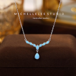 Op de afbeelding: Een zilveren ketting met een delicate ketting en een hanger met lichtblauwe opaalstenen. De ketting heeft een druppelvormige steen aan de onderkant en een reeks ovale stenen erboven. De tekst "MICHELLELEE STUDIO Handcrafted Jewellery" staat bovenaan.