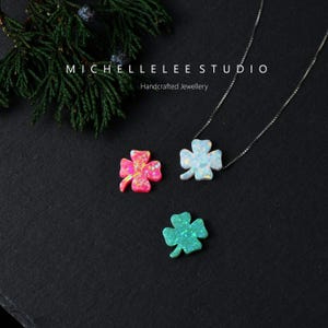 May include: Three opal-like clover pendants in pink, blue, and green. One pendant is on a silver chain. The pendants have a shimmering effect. The text "MICHELLELEELEE STUDIO Handcrafted Jewellery" is at the top.