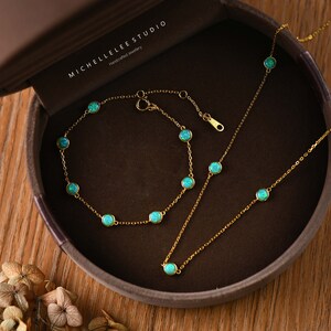 Pode incluir: Um conjunto de joias douradas com pulseira e colar, cada um adornado com pedras preciosas redondas azul claro. O conjunto &eacute; apresentado numa caixa de joias castanha. A marca "MICHELLELEE STUDIO" &eacute; vis&iacute;vel.