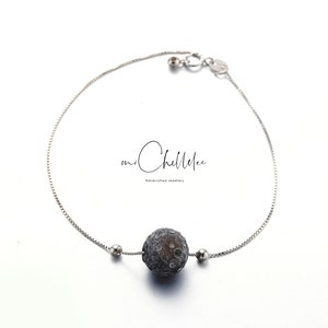 May include: A silver bracelet with a dark grey, textured sphere at its centre. Two small silver beads sit either side of the sphere. The bracelet has a fine chain and a clasp. The brand name "mi Chellelee" is written in cursive.