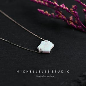 May include: A delicate silver chain necklace with a white opal pendant shaped like a house. The pendant has a textured surface and is set against a dark gray background.