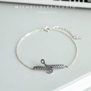 May include: A silver bracelet with an octopus charm. The octopus is detailed with small circles and has a textured surface. The bracelet is made of a delicate chain.