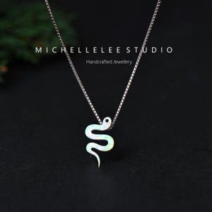 May include: A silver necklace featuring a snake pendant. The pendant is white with iridescent detailing. The necklace is set against a dark backdrop. The text "MICHELLELEE STUDIO Handcrafted Jewellery" is visible.