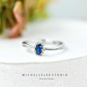 Op de afbeelding: Een zilveren ring met een blauwe ovale edelsteen, omringd door kleine heldere stenen en gouden accenten. De ring staat op een witte achtergrond. De tekst "MICHELLELEE STUDIO Handcrafted Jewellery" is zichtbaar.
