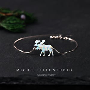 May include: A silver bracelet with an opal moose charm. The moose is white with a shimmering, multi-coloured effect. The bracelet has a fine chain and a lobster clasp. The text "MICHELLELEE STUDIO" and "Handcrafted Jewellery" are at the bottom.