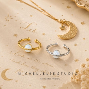 Könnte beinhalten: Eine goldene Halskette mit einem Anhänger in Form eines Halbmonds und eines Sterns sowie zwei offenen Ringen mit weißen, opalartigen Steinen. Ein Ring ist goldfarben, der andere silberfarben. Der Text "MICHELLE LEE STUDIO Handcrafted Jewellery" ist sichtbar.