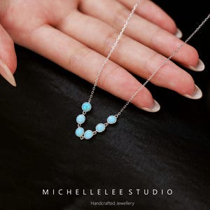 May include: A delicate silver necklace featuring a series of light blue gemstones. The gemstones are evenly spaced along the chain. The necklace is displayed against a dark background. The text "MICHELLELEE STUDIO" and "Handcrafted Jewellery" are visible.