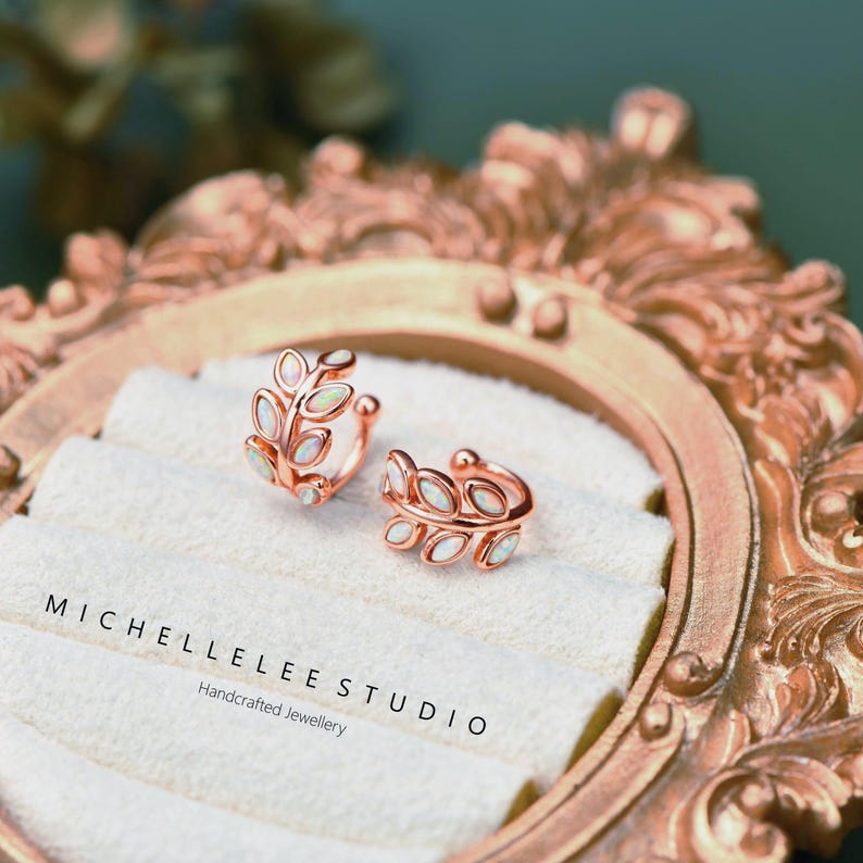 Puede incluir: Un par de pendientes tipo ear cuff de oro rosa con dise&ntilde;o de hojas y peque&ntilde;as piedras ovaladas iridiscentes. El texto MICHELLELEE STUDIO Handcrafted Jewellery est&aacute; impreso sobre la superficie de terciopelo blanco.