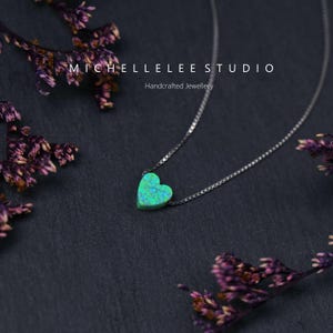 K&ouml;nnte beinhalten: Eine silberne Halskette mit einem herzf&ouml;rmigen Anh&auml;nger in Gr&uuml;n- und Blaut&ouml;nen. Die Kette wird vor einem dunklen Hintergrund mit getrockneten lila Blumen pr&auml;sentiert. Der Text "MICHELLELEE STUDIO Handcrafted Jewellery" ist sichtbar.