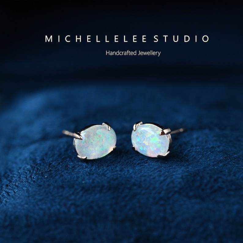 May include: A pair of silver stud earrings with oval-shaped white opal gemstones. The earrings are set against a blue velvet background.