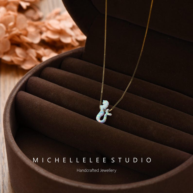Puede incluir: Un delicado collar de oro con un colgante en forma de ampersand. El colgante es blanco con detalles iridiscentes. El collar se muestra en un joyero marr&oacute;n. El texto "MICHELLELEE STUDIO Handcrafted Jewellery" es visible.