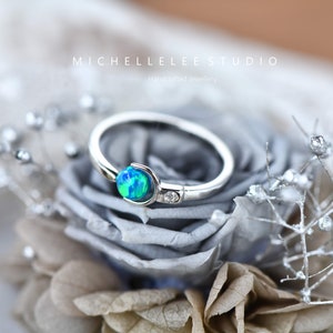 May include: A silver ring featuring a blue and green opal gemstone with a small diamond accent. The ring is displayed on a grey and white flower.