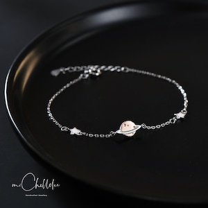 May include: A silver chain bracelet with a small planet charm and two star charms. The bracelet is on a black surface.