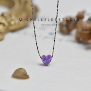 K&ouml;nnte beinhalten: Eine zarte Halskette mit einem kleinen, herzf&ouml;rmigen Anh&auml;nger in Lila- und Blaut&ouml;nen. Der Anh&auml;nger h&auml;ngt an einer feinen, dunklen Kette. Der Text "MICHELLELEE STUDIO Handcrafted Jewellery" ist sichtbar.