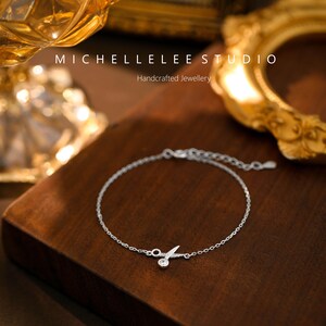 May include: A delicate silver bracelet with a small, clear crystal charm in the shape of a pair of scissors. The bracelet is on a wooden surface. Michellelee Studio Handcrafted Jewellery.