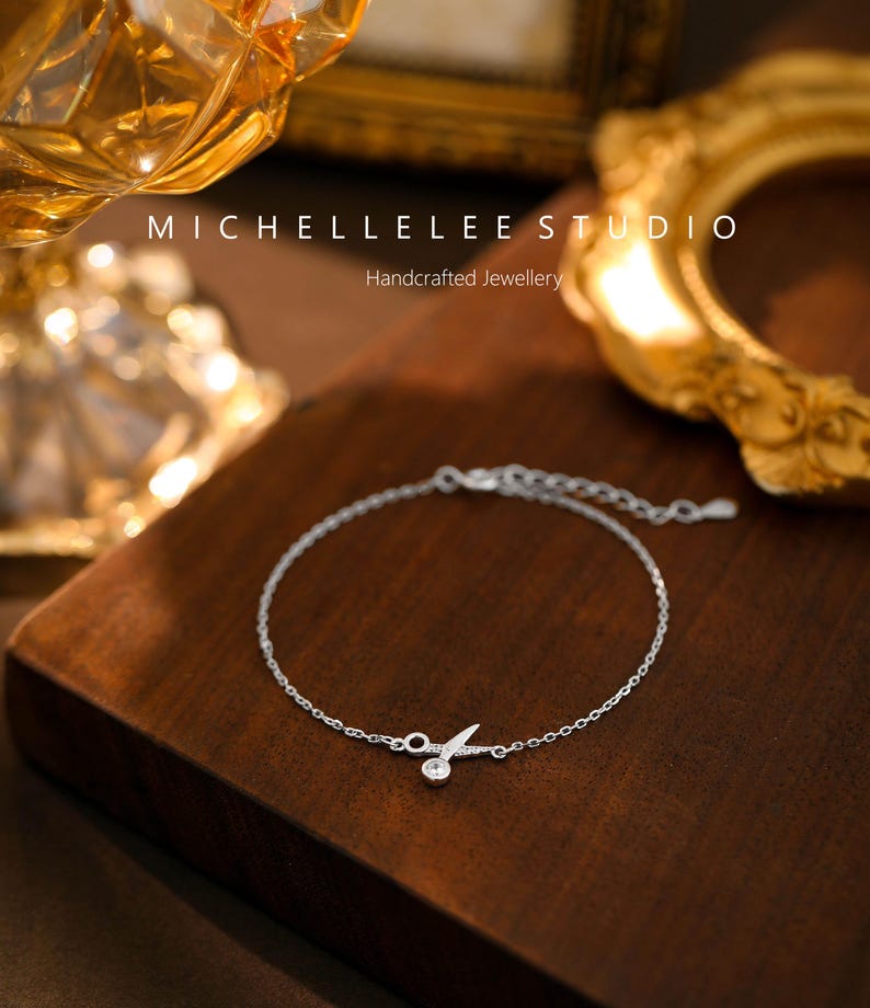 May include: A delicate silver bracelet with a small, clear crystal charm in the shape of a pair of scissors. The bracelet is on a wooden surface. Michellelee Studio Handcrafted Jewellery.