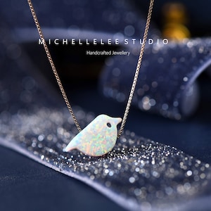 May include: A delicate silver chain necklace with a small, iridescent opal bird pendant. The bird is facing right and has a small black eye. The necklace is on a sparkly blue background.  Michelle Lee Studio, Handcrafted Jewellery.