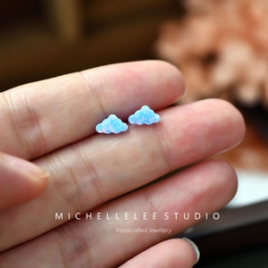 Pode incluir: Um par de pequenos brincos em forma de nuvem feitos de opala iridescente. Os brincos são de cor azul claro com um toque de rosa.