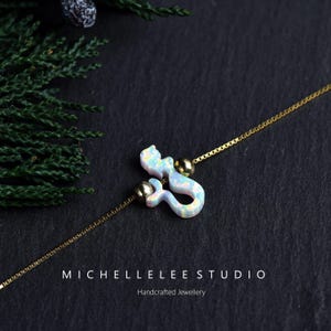 Puede incluir: Un collar de cadena dorada con un colgante de sirena blanco e iridiscente. El colgante se acent&uacute;a con dos cuentas doradas. El collar se exhibe sobre una superficie oscura con el texto "MICHELLELEE STUDIO" y "Handcrafted Jewellery".