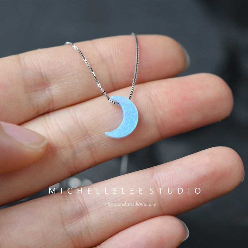 May include: A delicate silver necklace featuring a crescent moon pendant. The moon is a light blue opal, and the chain is a fine silver box chain. The necklace is a piece of handcrafted jewelry.