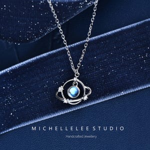 Könnte beinhalten: Eine silberfarbene Halskette mit einem planetenförmigen Anhänger, der einen zentralen schillernden blauen Stein enthält, umgeben von einem dünnen Orbitalring mit kleinen klaren Steinen. Der Text MICHELLELEE STUDIO Handcrafted Jewellery ist sichtbar.