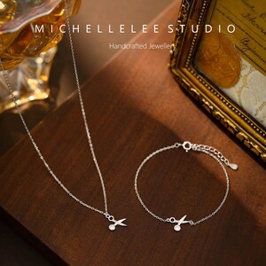 May include: A silver necklace and bracelet with a small scissor charm. The jewelry is on a wooden surface. The text "MICHELLELEE STUDIO Handcrafted Jewellery" is visible in the background.